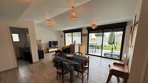 Un vaste espace de vie comprenant une table à manger, des chaises, un téléviseur et une porte-fenêtre coulissante donnant sur une terrasse extérieure.