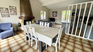 Un espace repas comprenant une grande table et des chaises, une cheminée, un téléviseur et un coin cuisine visible à travers une porte ouverte.