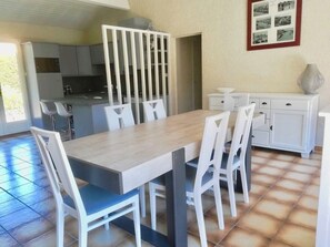 Un coin repas avec une table en bois et des chaises blanches, une cuisine en arrière-plan et un sol carrelé.
