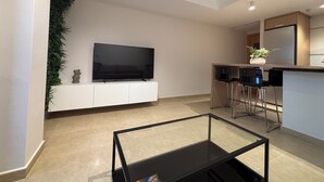 En moderne stue med et fladskærms-tv, et glasbord og et køkkenområde med et køleskab og barstole.