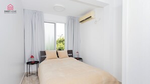 A modern bedroom with a large bed, two bedside tables with lamps, a window with curtains, and an air conditioning unit. - Quiet Oasis in the Center (Netanya)