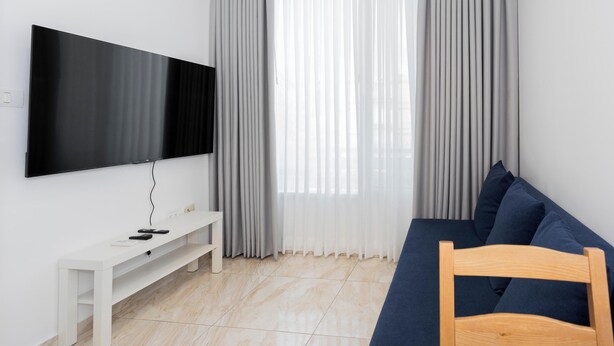 A modern living room with a flat-screen TV mounted on the wall, a white bench, a dark blue sofa, and a wooden chair. - Quiet Oasis in the Center (Netanya)