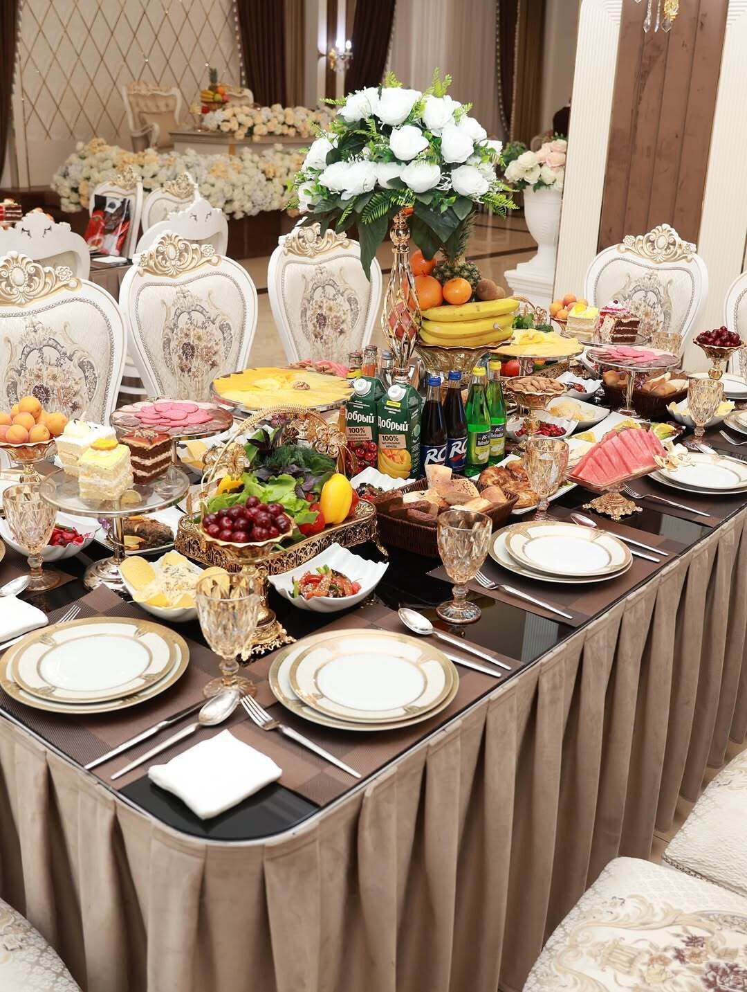 Una mesa de banquete dispuesta con diversos platos, frutas y bebidas, rodeada de sillas ornamentadas.