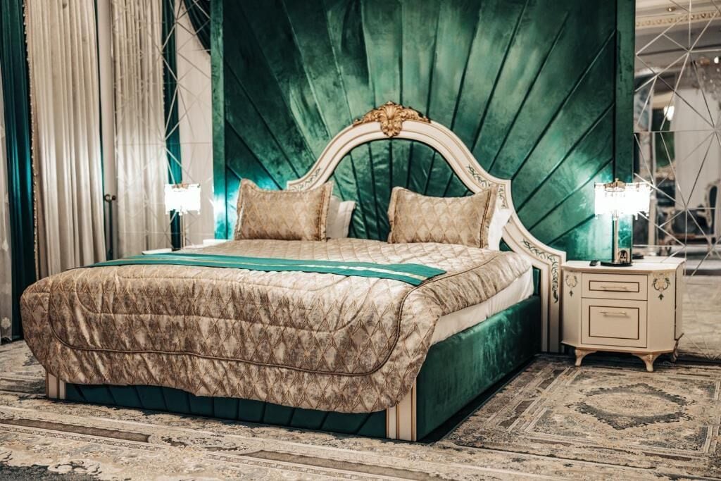 Una cama grande con cabecera tapizada en verde, dos lámparas de noche y una colcha estampada.