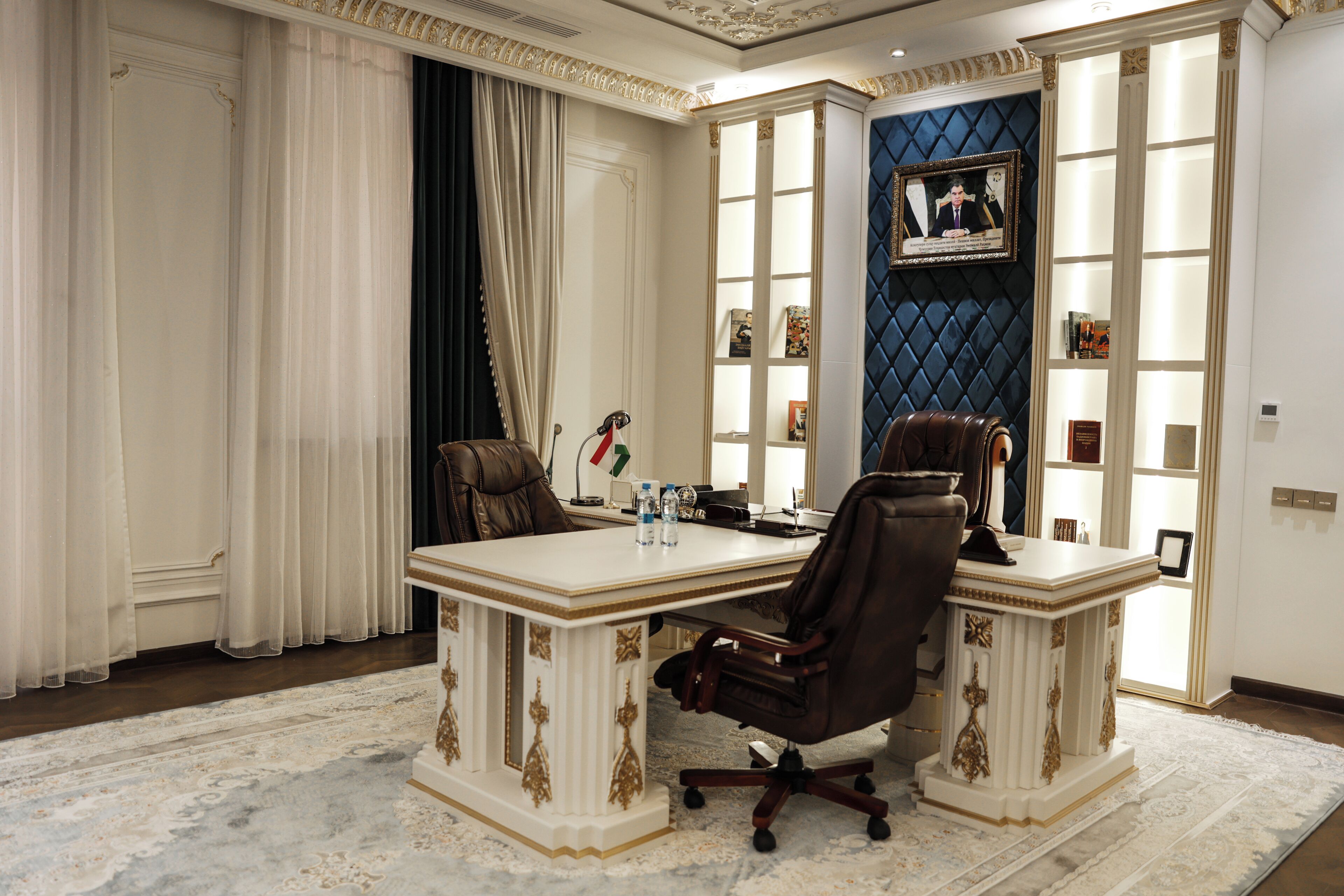 A formal meeting room with a large table, two chairs, a bookshelf, and a framed portrait on the wall.