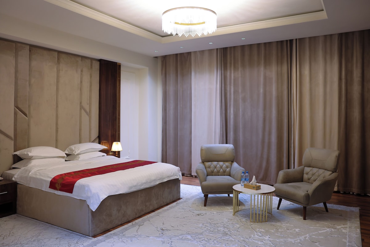 Una habitación de hotel con una cama grande, dos sillones, una mesita redonda y una araña.