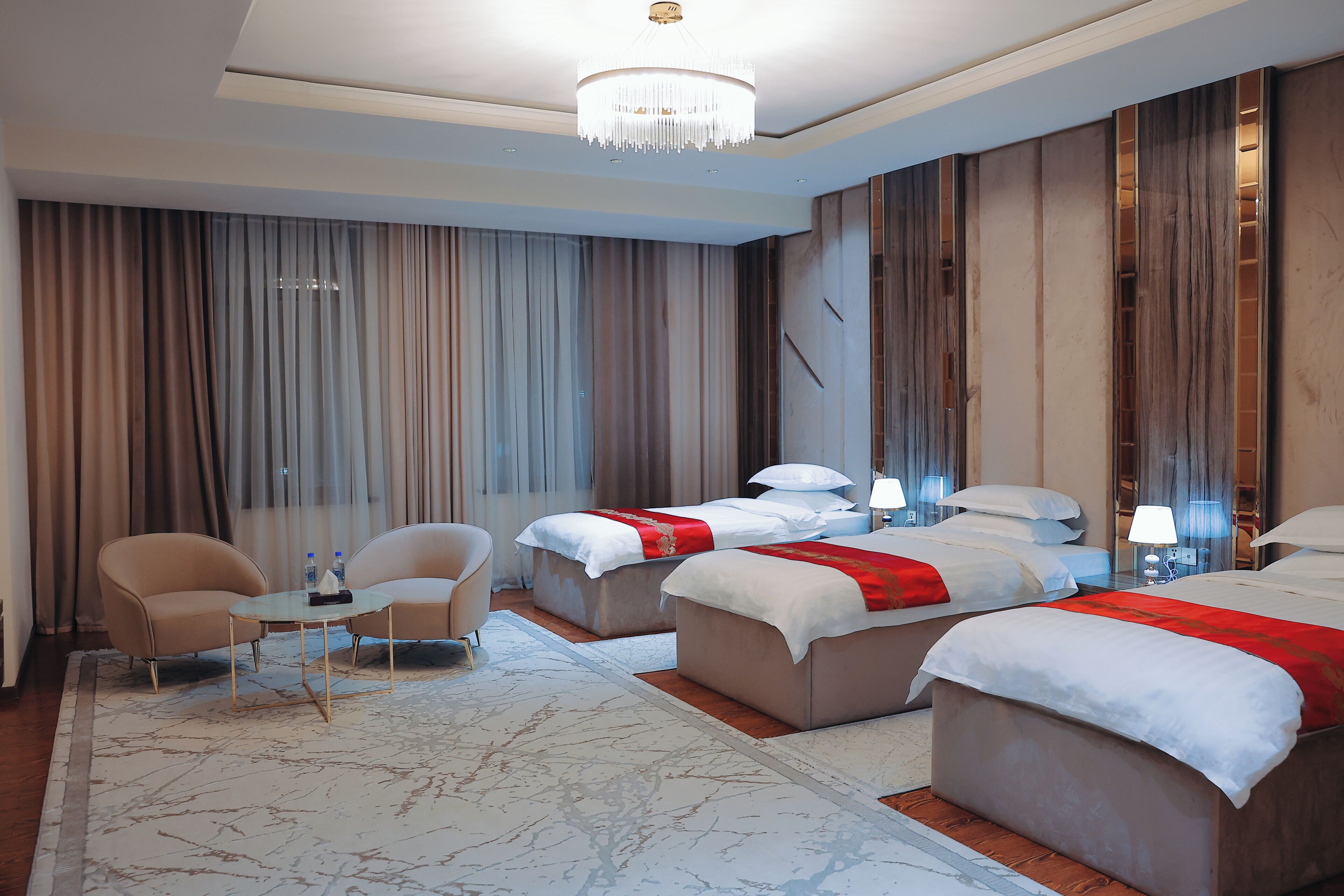Habitación de hotel con dos camas, un área de descanso con sillones, una araña y un amplio ventanal con cortinas.