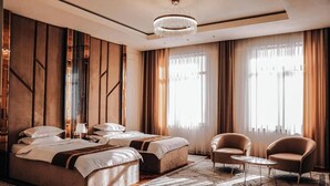 Ένα σύγχρονο δωμάτιο ξενοδοχείου με ένα μεγάλο κρεβάτι, δύο κομοδίνα, ένα πολυέλαιο και μεγάλα παράθυρα με κουρτίνες.