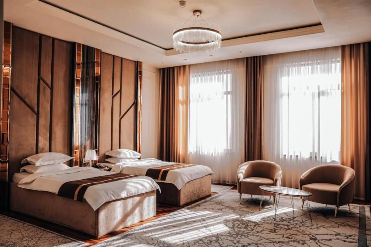 Una habitación de hotel moderna con una cama grande, dos mesitas de noche, un candelabro y ventanas amplias con cortinas.