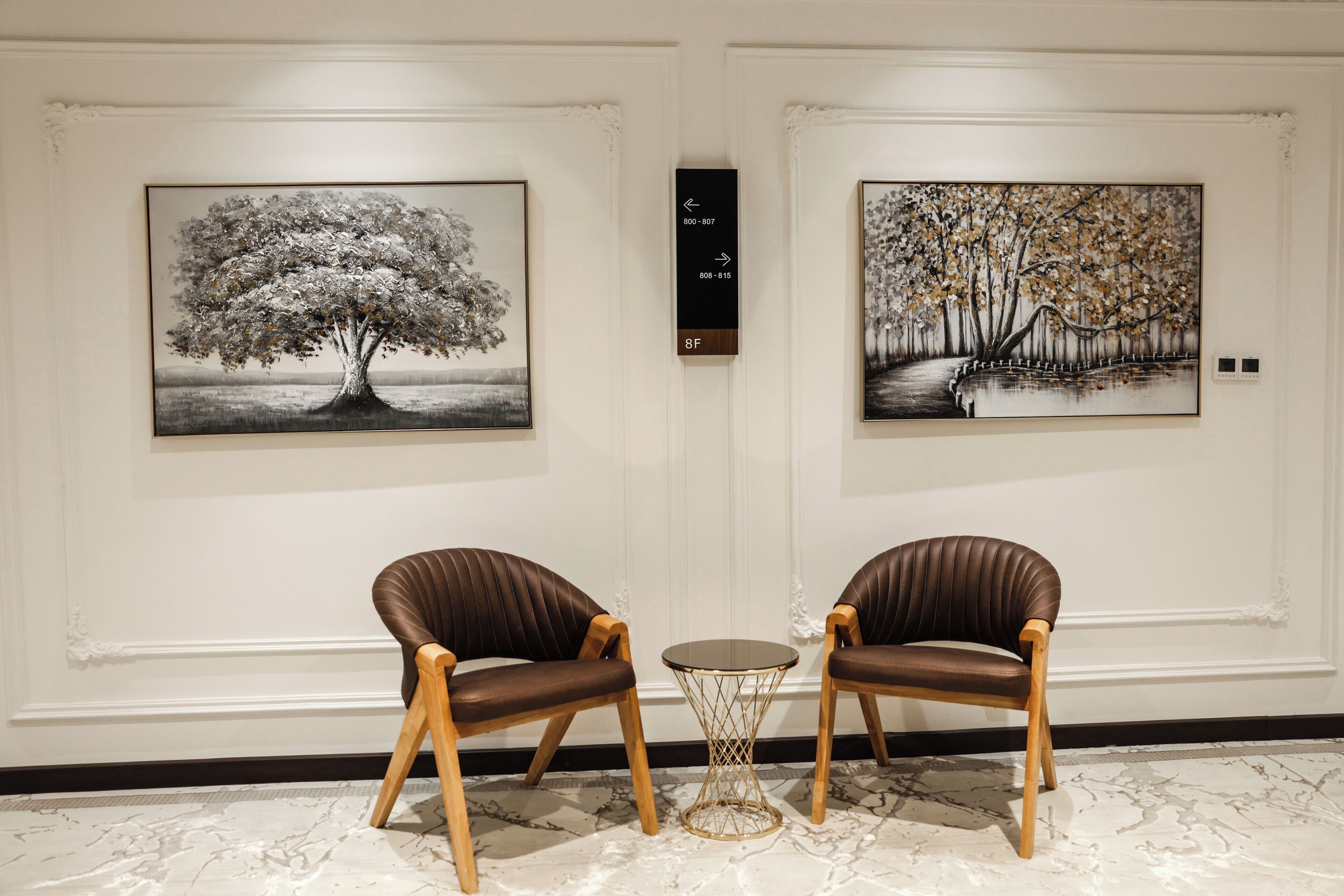 Two leather chairs facing a small round table with a wireframe base, positioned between two framed paintings of trees.