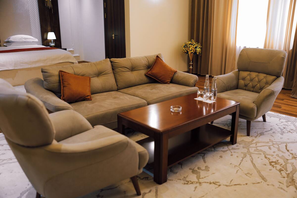 Ein Hotelzimmer mit einem großen, beigen Sofa, zwei Sesseln, einem Holztisch und einem Bett mit weißer Bettdecke.