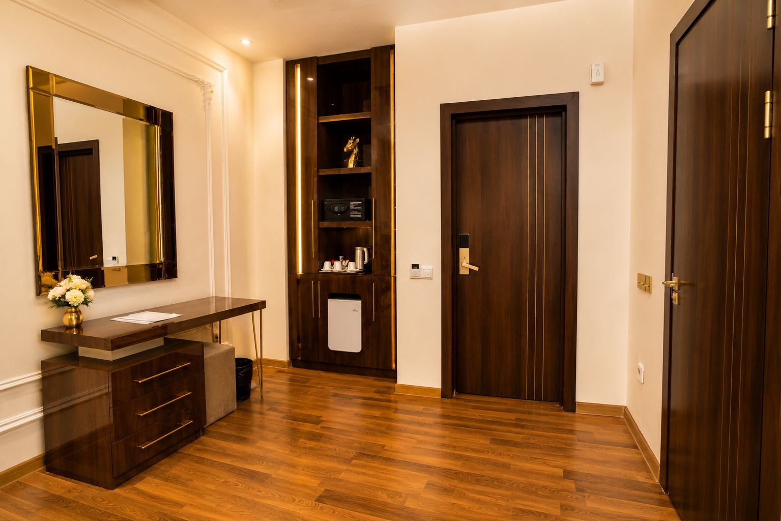 A hotel room with a wooden floor, a dark wood vanity with a mirror, a small refrigerator, and a door with a handle.