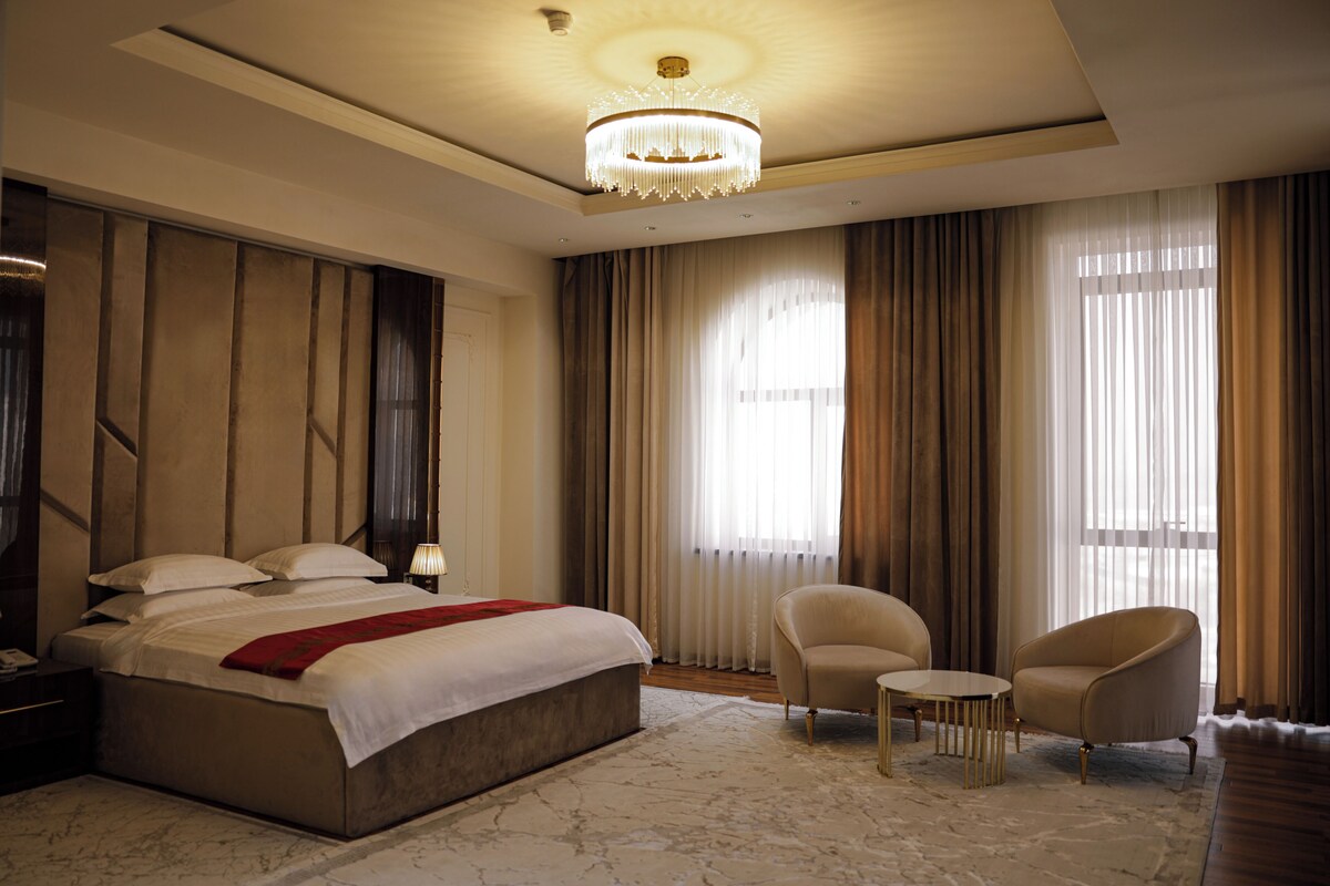 A spacious bedroom with a large bed, two armchairs, a small table, and a chandelier.