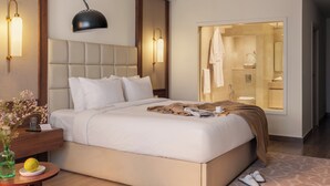 Ein modernes Hotelzimmer mit einem großen Bett, Nachttischlampen, einem Nachttisch mit einer Vase und Obst, sowie Blick ins Badezimmer.
