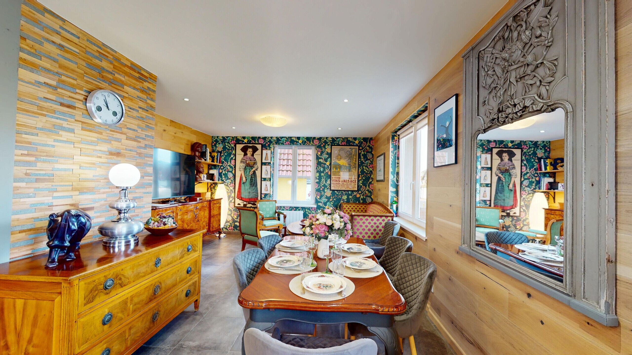 Un comedor con una mesa de madera dispuesta para cuatro personas, rodeada de sillas de estilo vintage. Un televisor está montado en la pared, y un espejo grande refleja la habitación.