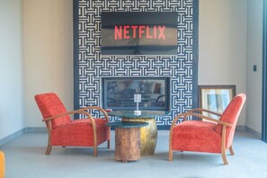 Un espace de vie moderne avec deux fauteuils rouges à revêtement en tissu, une table basse en verre et un téléviseur fixé au mur qui affiche le logo « NETFLIX ».