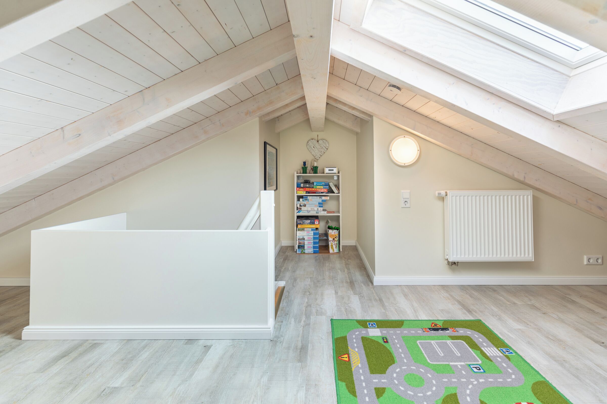 Une pièce mansardée aux hauts plafonds, dotée d’une lucarne, d’un banc intégré blanc, d’un tapis de jeu pour enfants et d’une bibliothèque remplie de livres et de jouets.