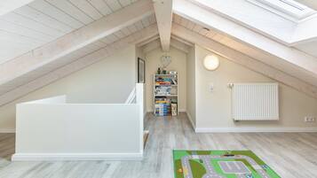 Une pièce mansardée aux hauts plafonds, dotée d’une lucarne, d’un banc intégré blanc, d’un tapis de jeu pour enfants et d’une bibliothèque remplie de livres et de jouets.