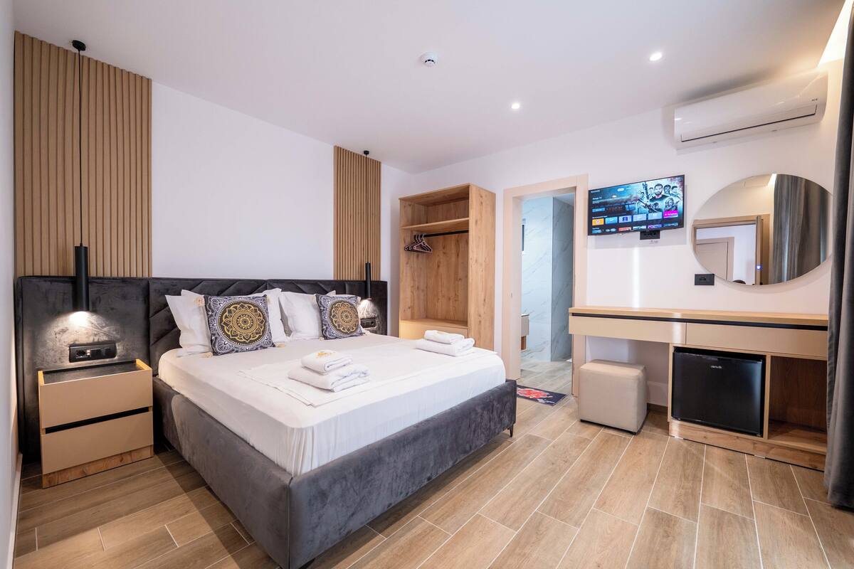 Una habitación de hotel moderna con una cama grande, un armario de madera, un escritorio empotrado, un televisor y un refrigerador compacto.