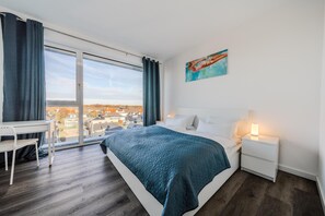 A bedroom with a bed, a desk, a chair, a nightstand, a lamp, and a painting. - Loft Fluxus Emden | Delft View (Emden)