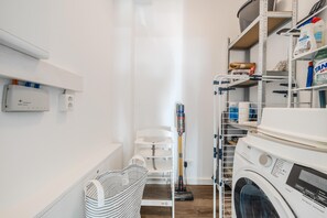A laundry room with a front-loading washing machine, shelves with cleaning supplies, a vacuum cleaner, and a white chair. - Loft Fluxus Emden | Delft View (Emden)