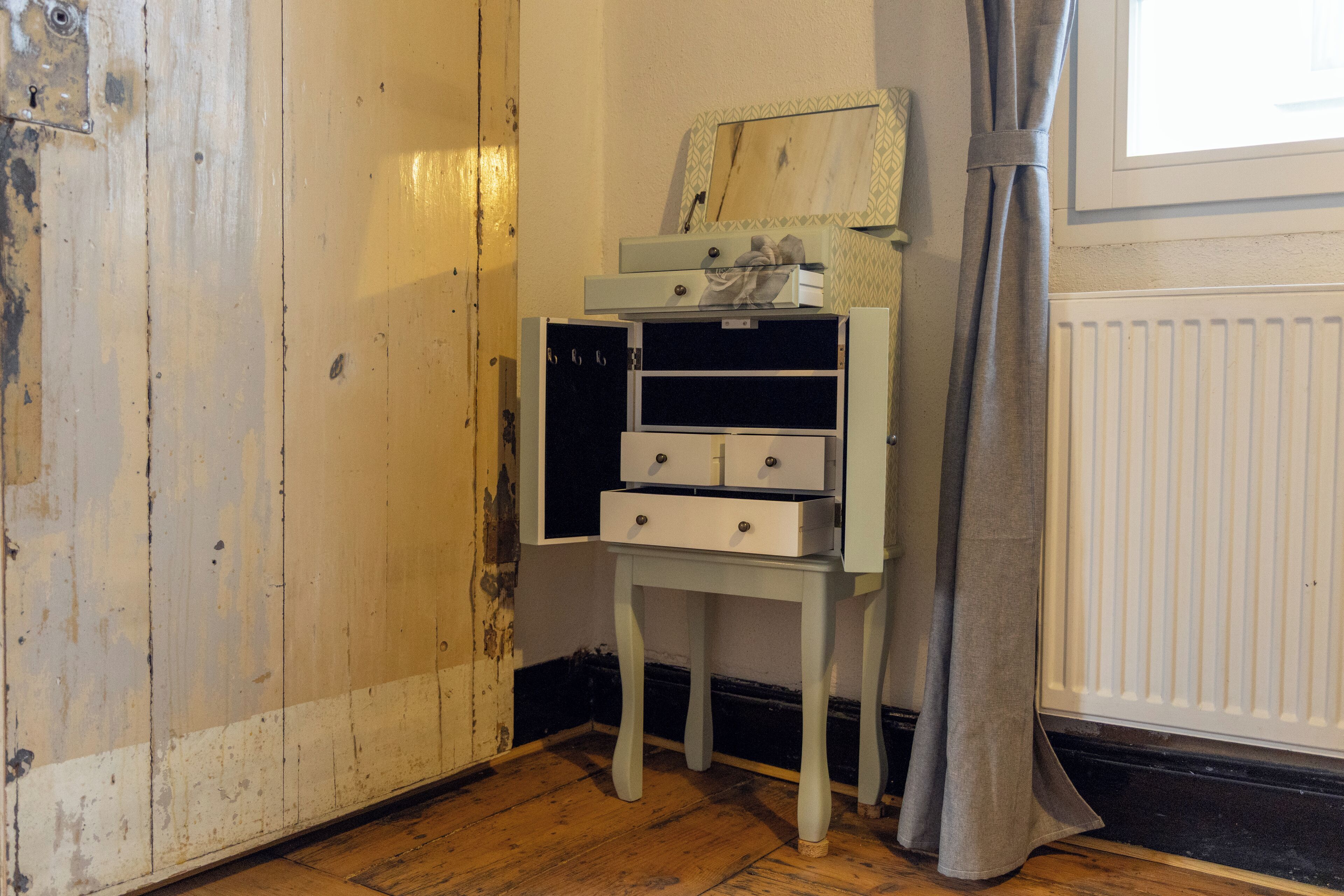 A room with a wooden floor, a white radiator, a grey curtain, and a small white table with drawers.