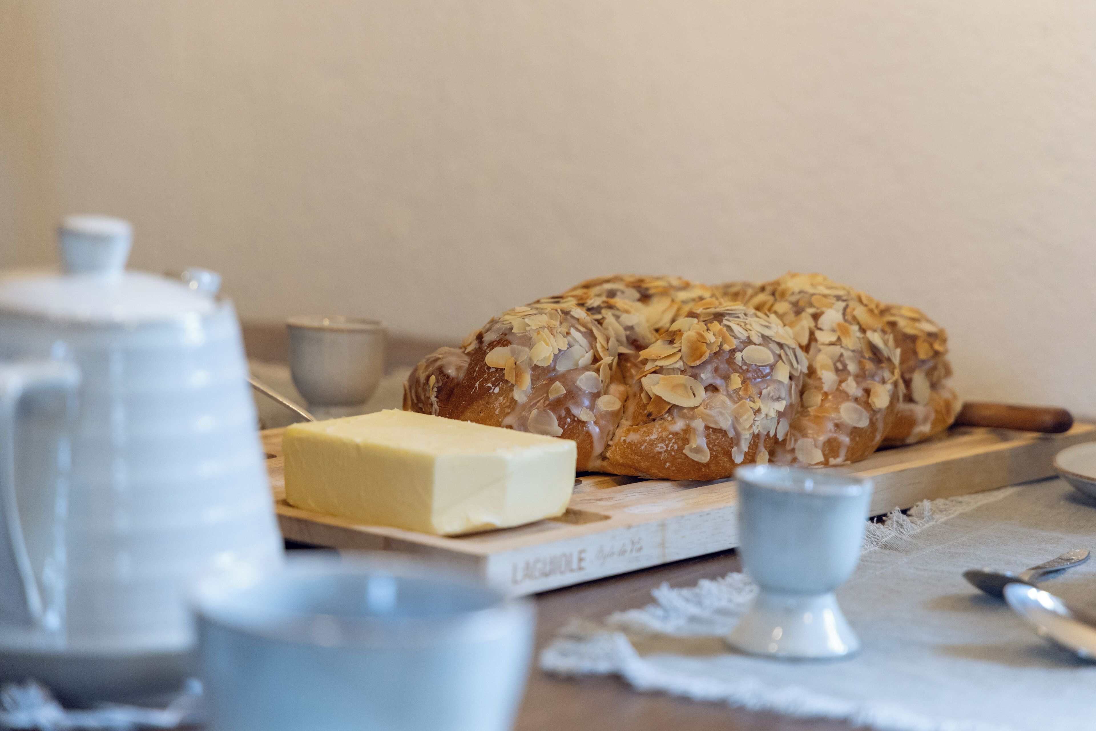 Leikattu leipä, jossa on manteleita, on asetettu puisen leikkuulaudan päälle. Sen vieressä on voinokare ja pieni kannu pöydällä.