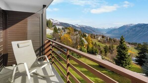 Un balcón con una silla blanca, baranda de madera y vistas a un paisaje montañoso con edificios a lo lejos.