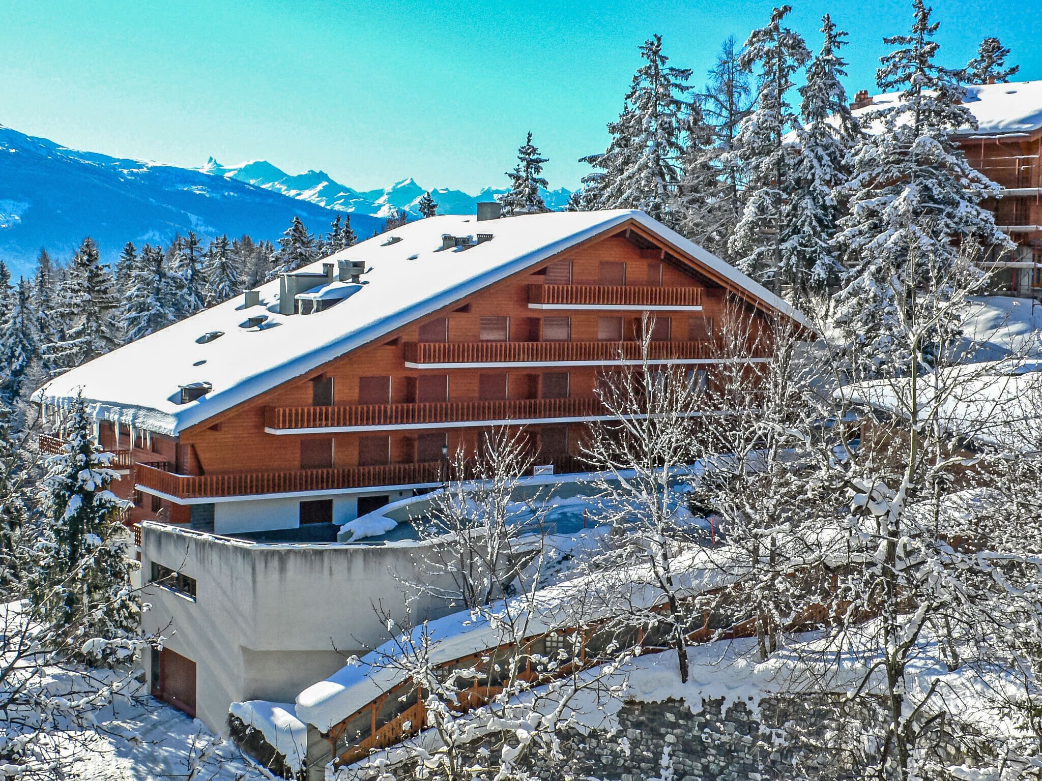 Ein modernes Skiresort-Gebäude mit mehreren Balkonen, umgeben von schneebedeckten Bäumen und Bergen im Hintergrund.