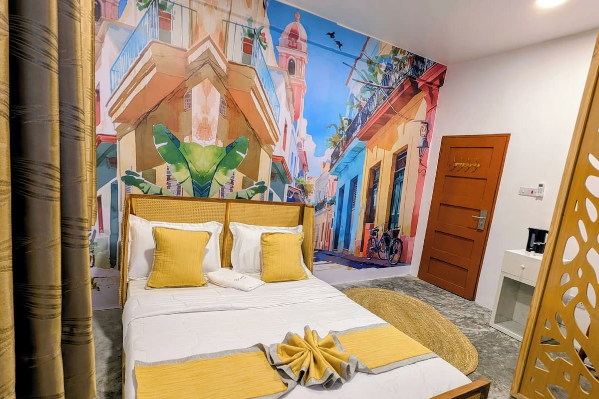A hotel room with a bed, a mural of a street scene, a wooden door, and a small desk with a lamp.