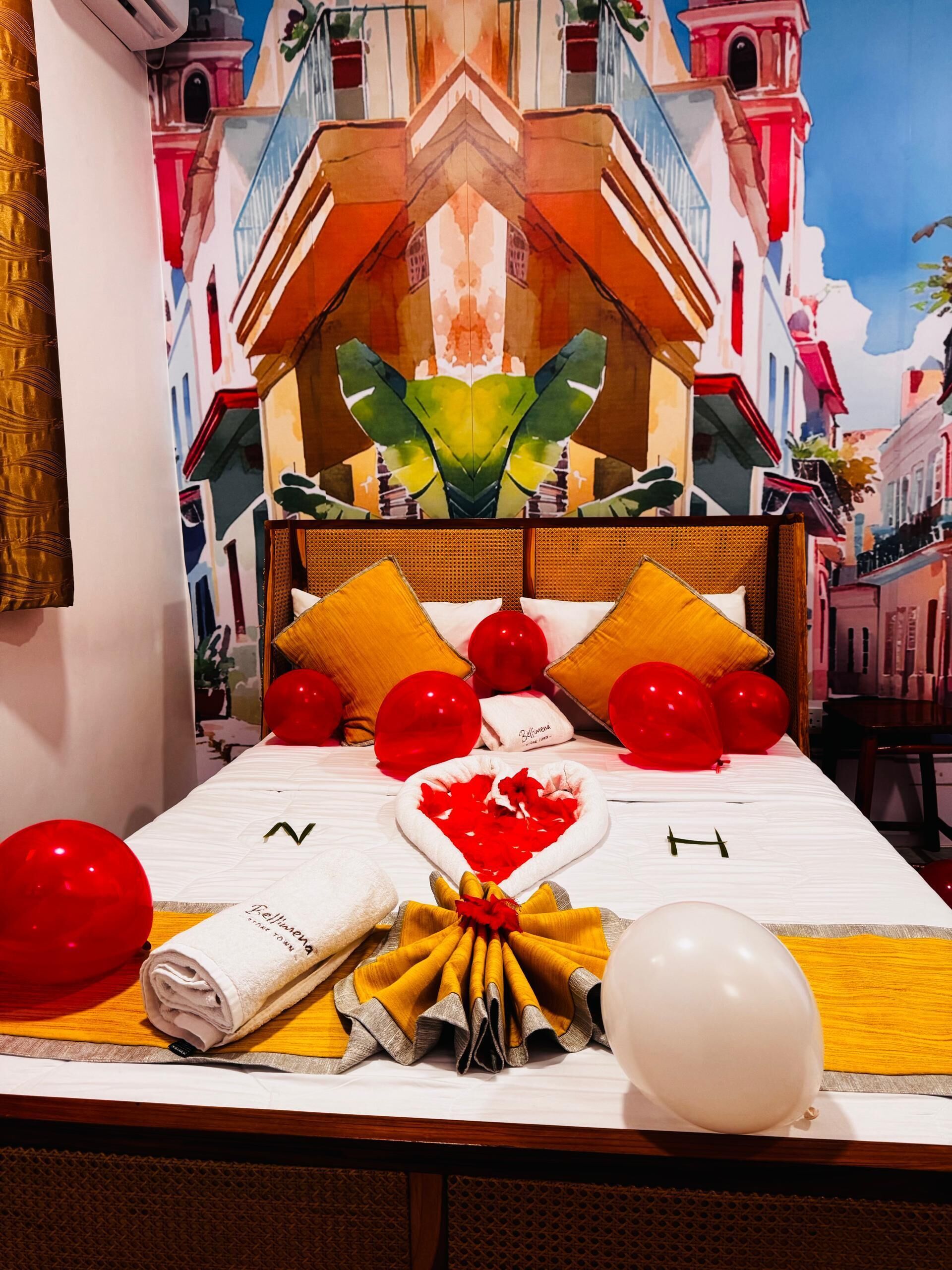 A bed with a heart-shaped pillow, red and white balloons, and a towel with 'Bellissima' text.