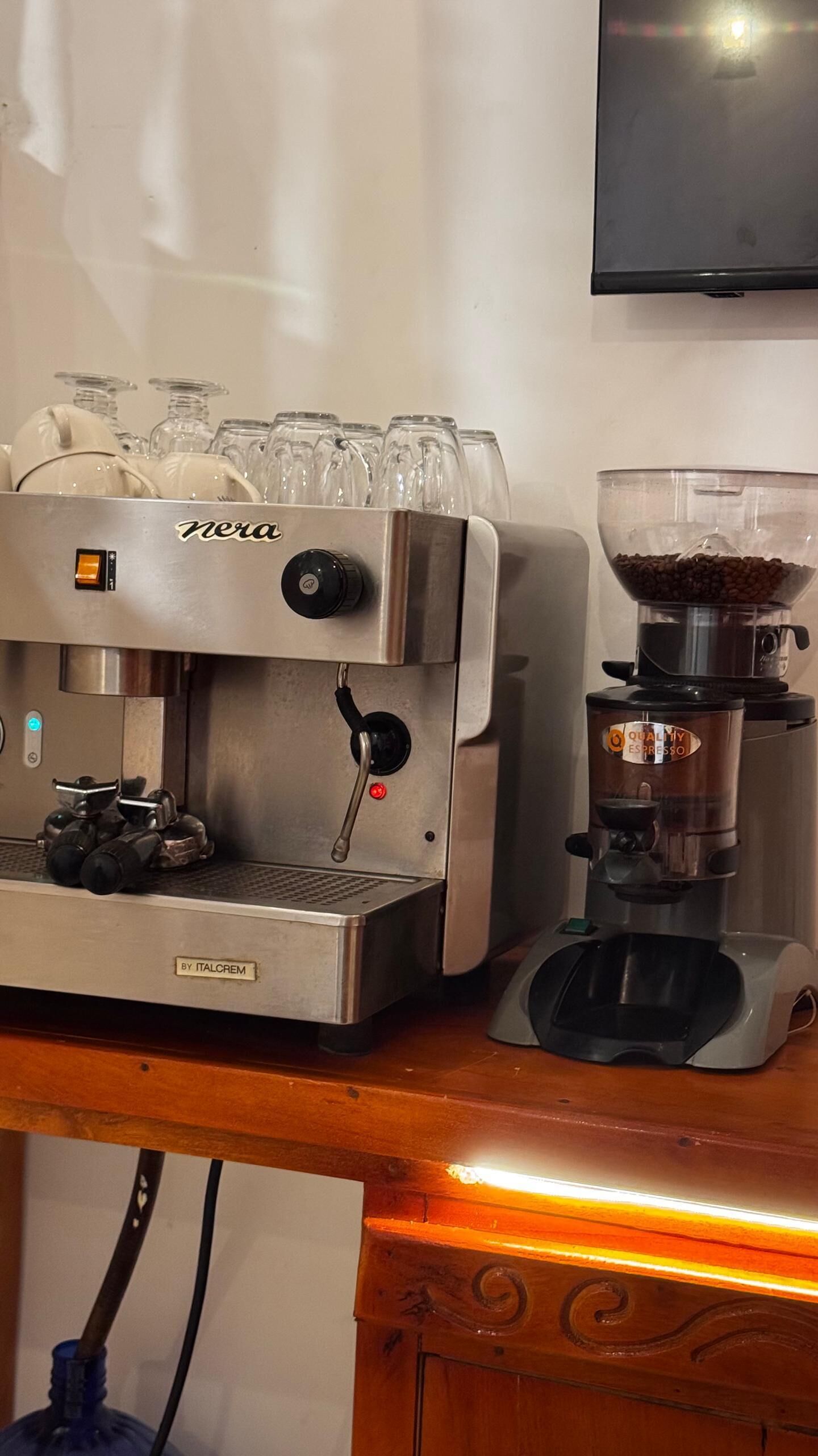 Una cafetera con un molinillo, una jarra de café, vasos y un televisor en la pared.