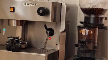 Una cafetera con un molinillo, una jarra de café, vasos y un televisor en la pared.