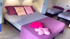 Ein Zimmer mit zwei Betten, jeweils mit gemustertem Bettbezug und Kissen. Ein kleiner Tisch mit rosa Tischdecke und mehrere rosafarbene Kissen sind vorhanden. Eine dekorative Vase mit Blumen steht auf einem Regal über den Betten.