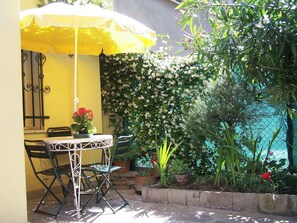 Une terrasse avec un parasol jaune, une table et des chaises. Un mur est recouvert de plantes grimpantes, et on peut admirer une étendue d’eau.