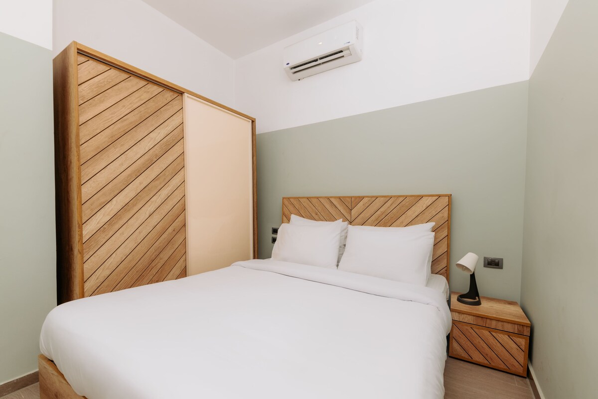 Un dormitorio con cabecera de madera, cama con ropa de cama blanca, mesita de noche con lámpara y un equipo de aire acondicionado en la pared.