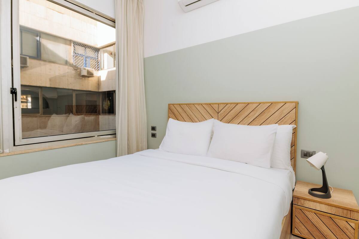 Una cama bien hecha con sábanas blancas, cabecera de madera y una lámpara de noche sobre una mesita de madera.