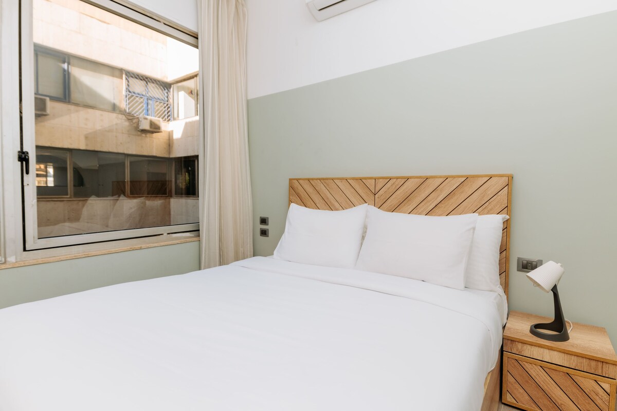 Habitación de hotel con cama, mesita de noche con lámpara, ventana con cortinas y vista a un edificio.