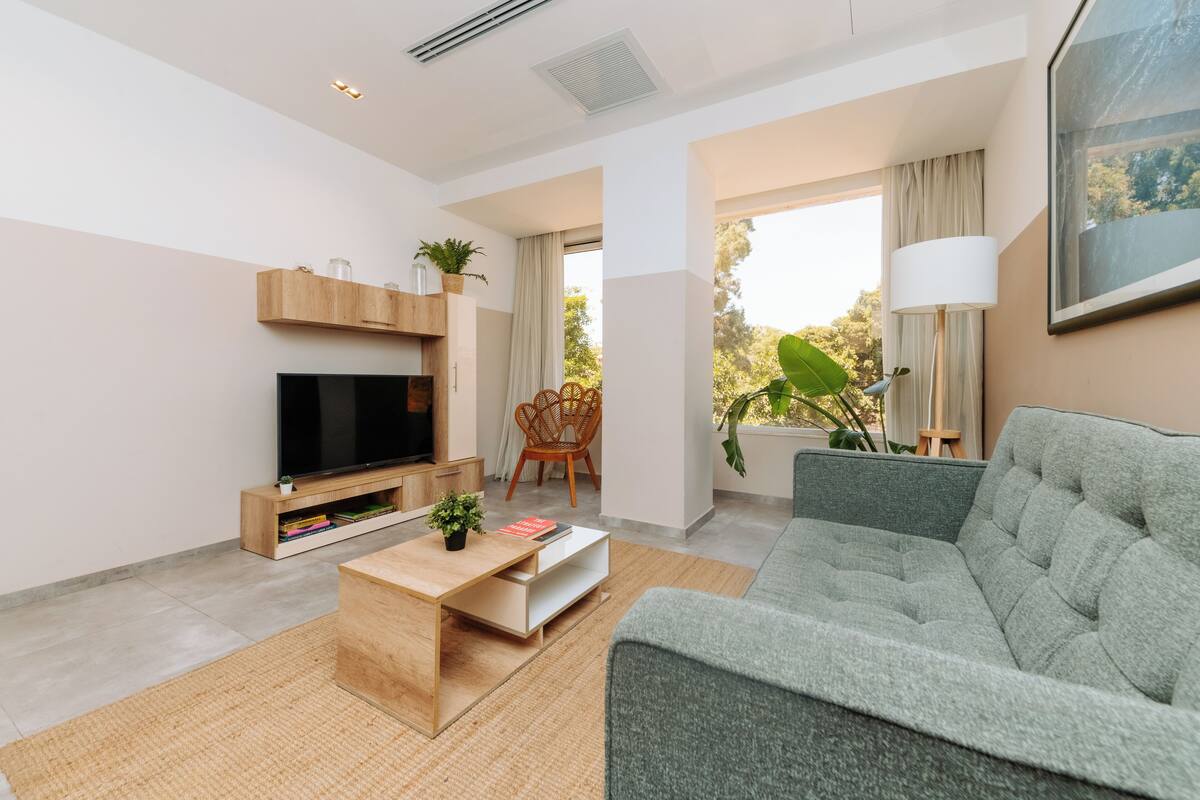 Un salón moderno con un televisor de pantalla plana, un sofá gris, una mesa de centro de madera y un amplio ventanal con cortinas.