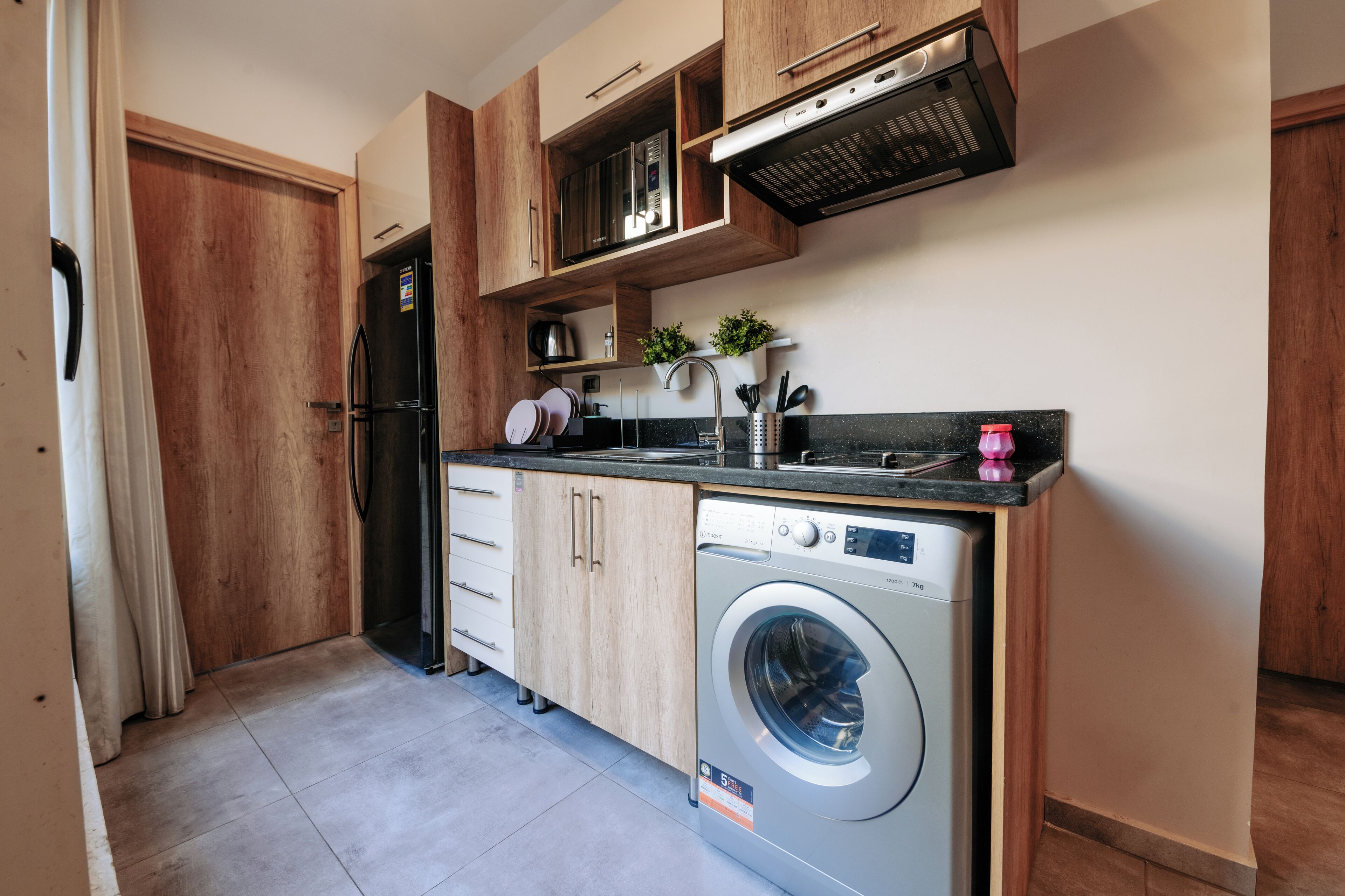 A compact kitchen with wooden cabinets, a built-in microwave, a stainless steel refrigerator, a washing machine, and a small countertop with a sink and a potted plant.
