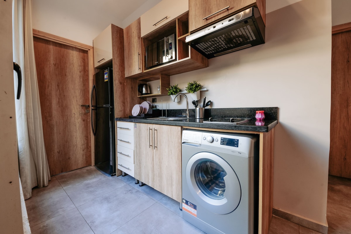 A compact kitchen with wooden cabinets, a built-in microwave, a stainless steel refrigerator, and a front-loading washing machine.