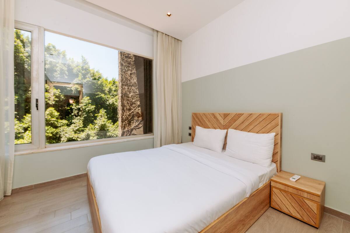 Un dormitorio con un ventanal grande, una cama con ropa de cama blanca, un cabecero de madera y una mesita de noche con un teléfono.