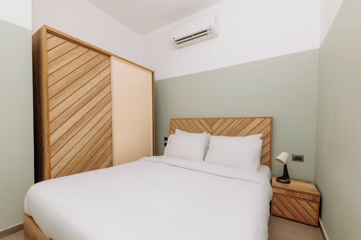 Una cama bien hecha con cabecera de madera, una mesita de noche con lámpara y un armario con patrón de espiga.