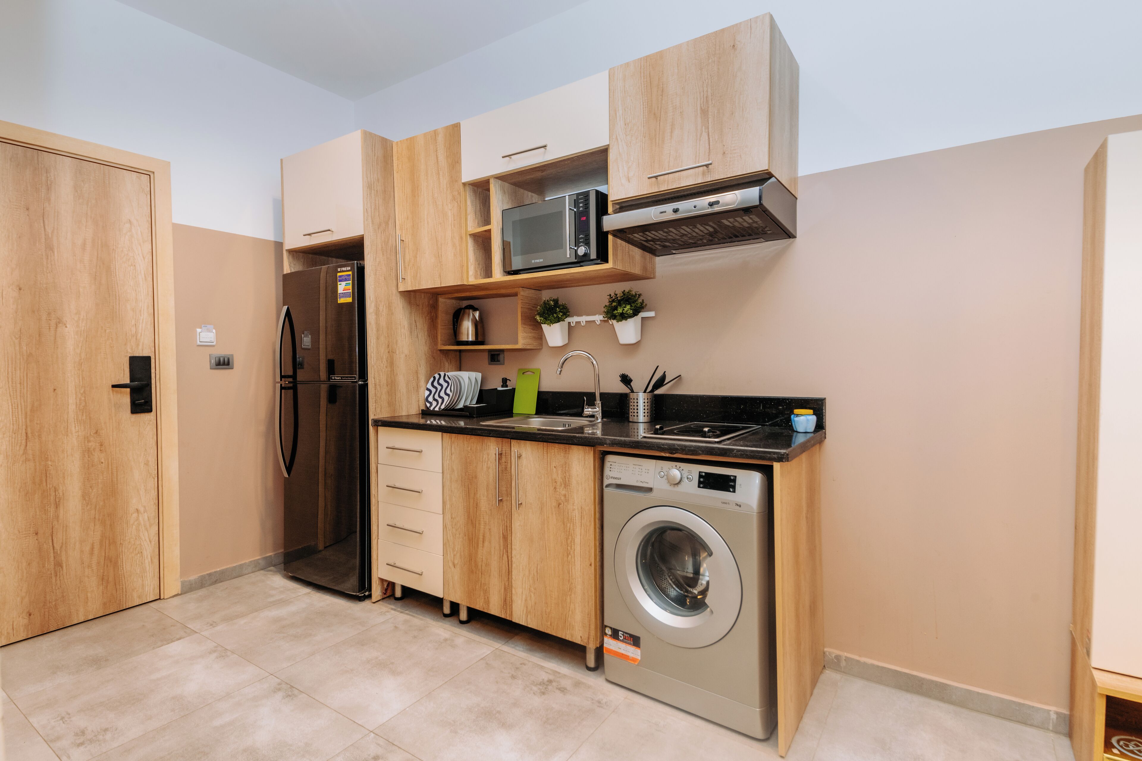 Una cocina compacta con armarios de madera, microondas integrado, refrigerador de acero inoxidable, lavadora y una pequeña encimera con fregadero y plantas en macetas.