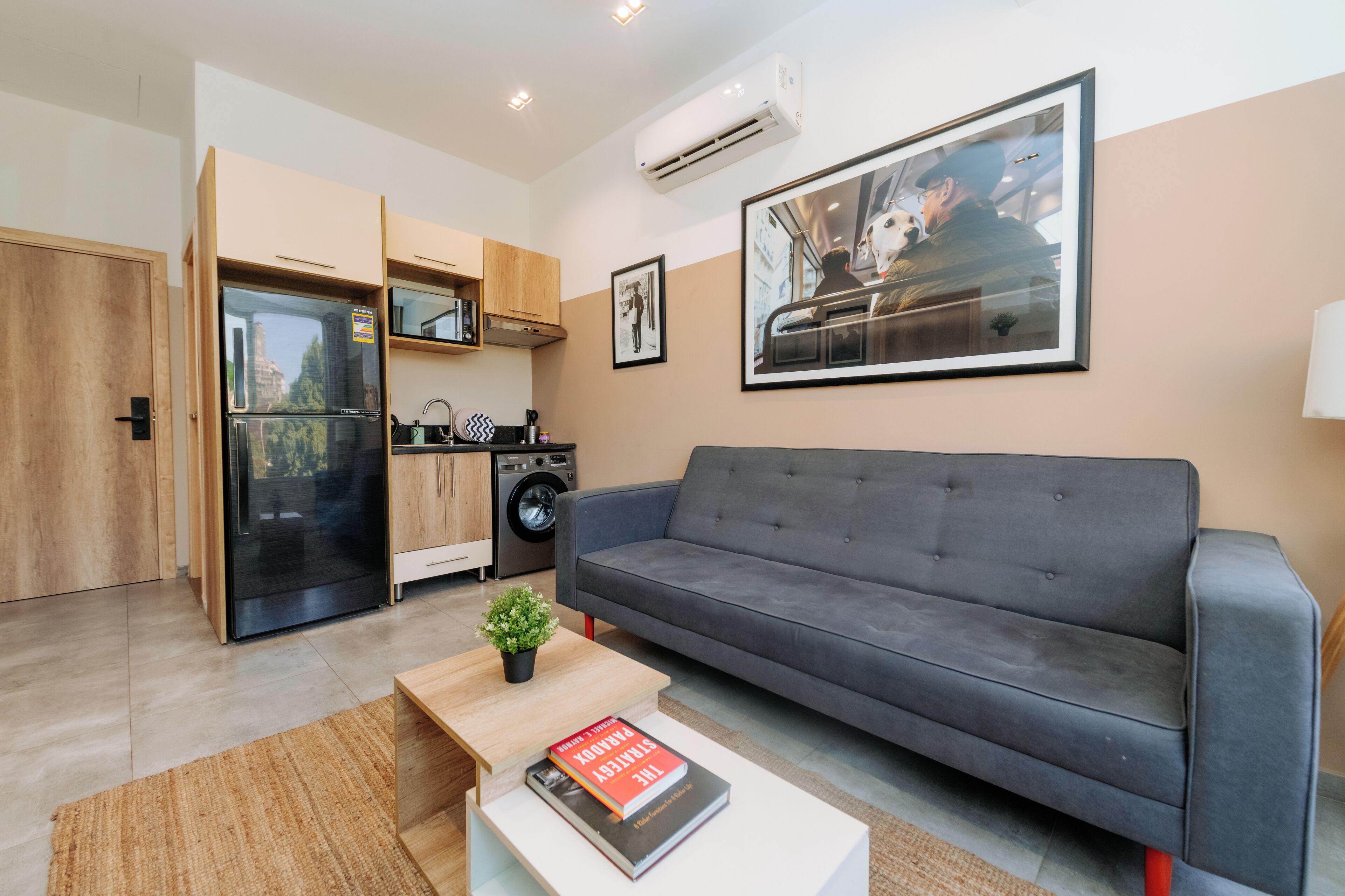 Una sala moderna con un sofá gris, una mesa de centro de madera y una cocina con lavadora y refrigerador.