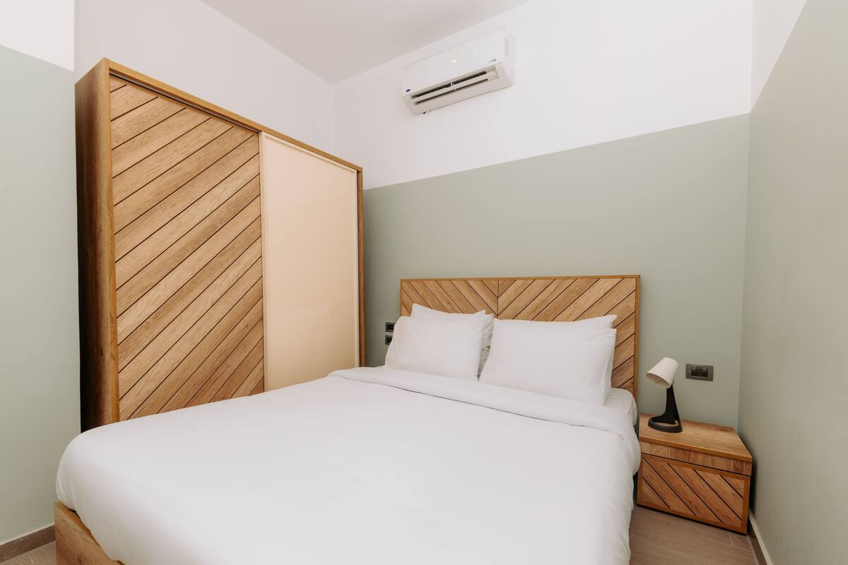 Un dormitorio con cabecera de madera, cama con ropa de cama blanca, mesita de noche con lámpara y un equipo de aire acondicionado en la pared.