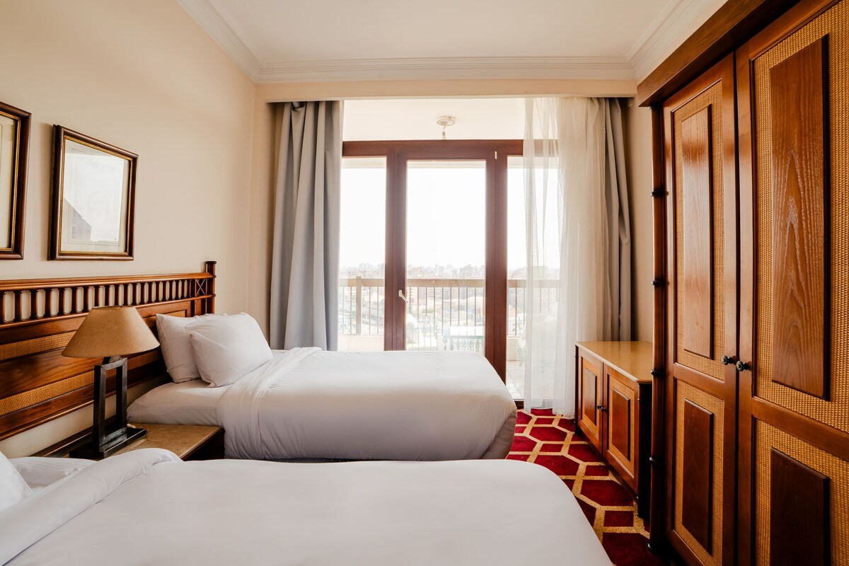 Une chambre d’hôtel avec deux lits, une armoire en bois, un bureau avec une lampe et une vue sur la ville grâce à de grandes fenêtres.