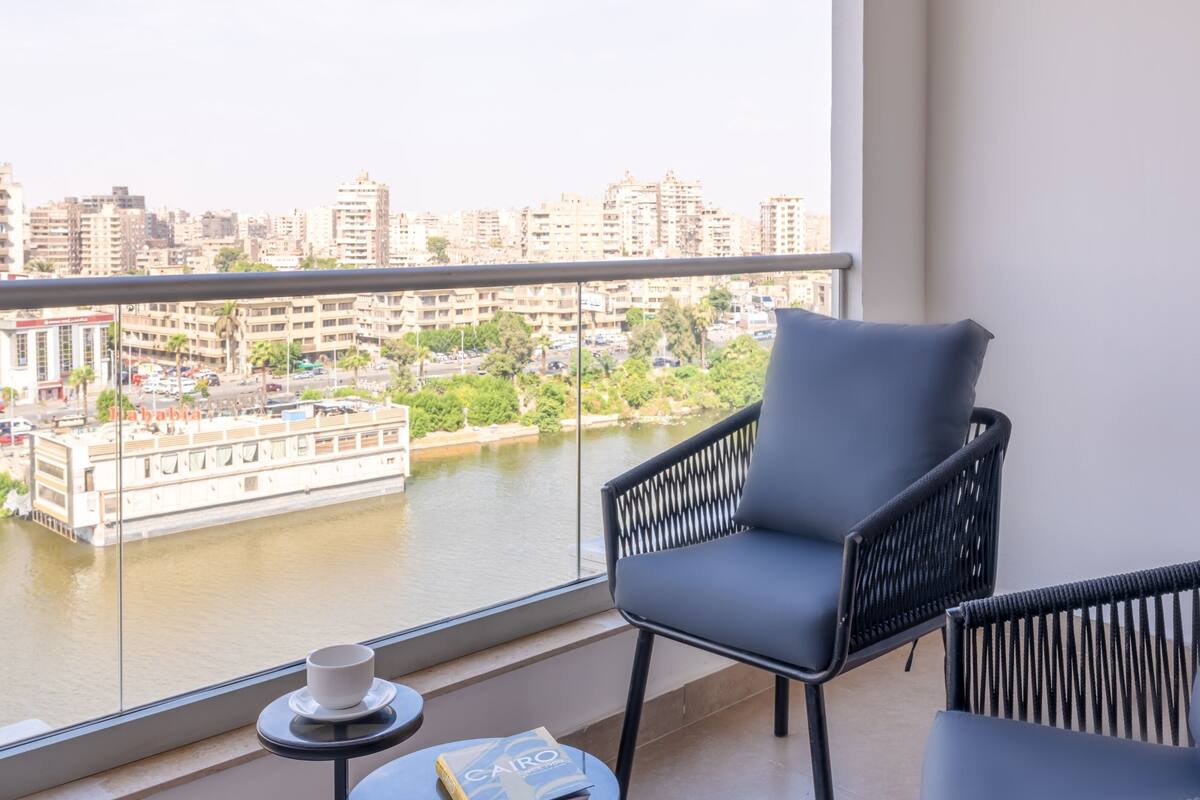 Un balcon donnant sur une rivière et des immeubles, une chaise, une table avec une tasse et un livre.