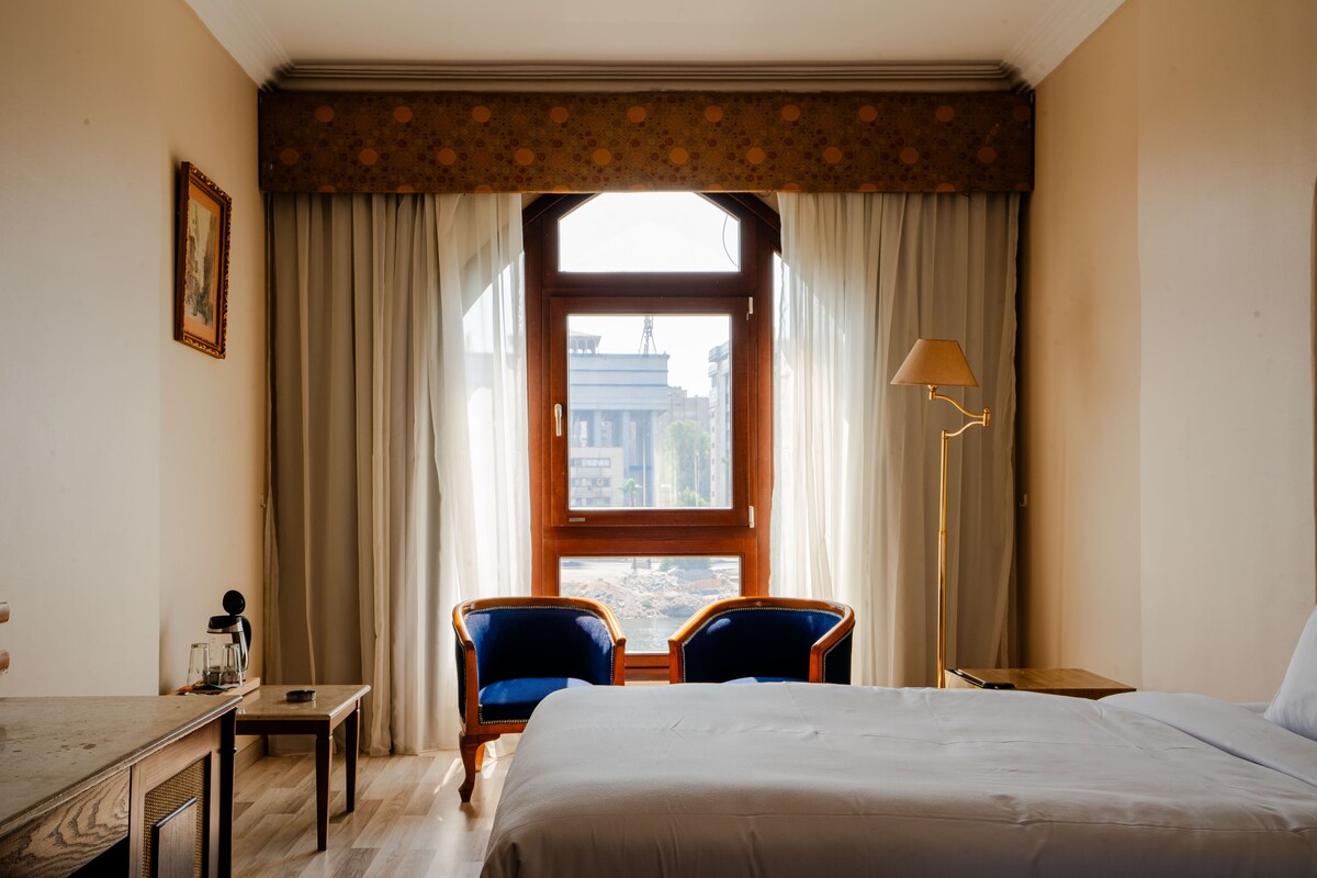Een hotelkamer met een bed, twee stoelen, een bureau, een lamp en een raam met gordijnen.
