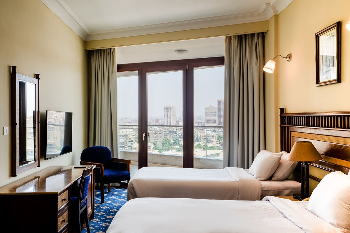 A hotel room with two beds, a desk, a chair, a lamp, and a large window with a city view.